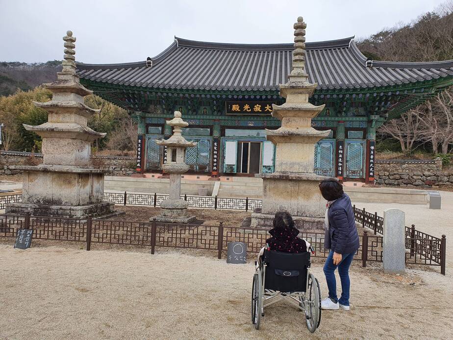 정면에 절이 보이고 양쪽으로 탑이 있는 사찰 마당에 휠체어를 타신 어르신이 있고 옆에는 중년 여성이 손으로 절을 가리키며 서 있다. 